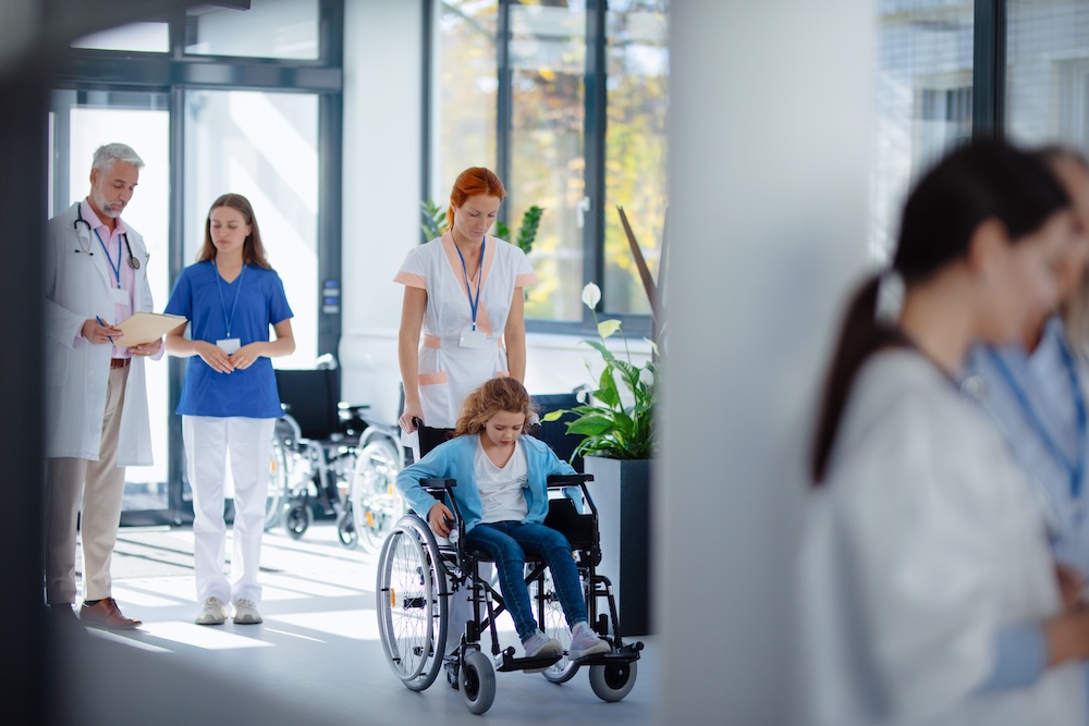 Young nurse pushing little girl on wheelchair at hospital corridor.